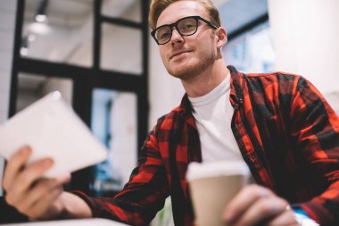 Gündelik giysiler ve gözlüklerle yakışıklı genç adam kahve dükkanında kameraya bakarak bir kupa taze içecek ve telefon alıyor.