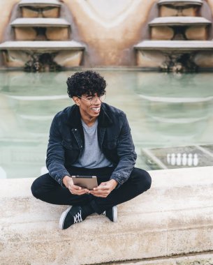 Genç, Afro-Amerikalı kıvırcık saçlı, fıskiyenin yanında bacak bacak üstüne atmış, elinde tabletle oturmuş başka tarafa bakan bir adam.