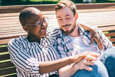 Neşeli, şefkatli, çok ırklı bir çift. Geniş ahşap bankta oturup sarılıyorlar. Güneşin tadını çıkarıyorlar.