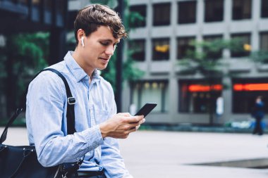 Resmi tişörtlü genç erkek işçi beton çitin üzerinde deri çantayla oturuyor ve New York 'un arka planında telefonla mesajlaşıyor.