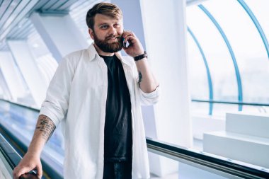 Elinde siyah tişört ve beyaz gömlekli şık sakallı adam cep telefonuyla konuşuyor hafif geometrik tünelde tırabzan tutuyor.