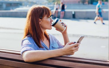 Rahat giyinen, arkadaşlarıyla telefon görüşmeleri yapan, sıcak içecek içen, limandaki ahşap bankta oturan sakin genç bayanın arka plan görüntüsü.