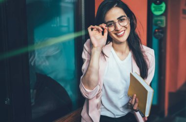 Yüksek açılı, zarif elbiseli, not defteri takan, gözlüklü ve şehirde ahşap bankta dinlenirken kameraya gülümseyen hoş bir yönetici kadın.