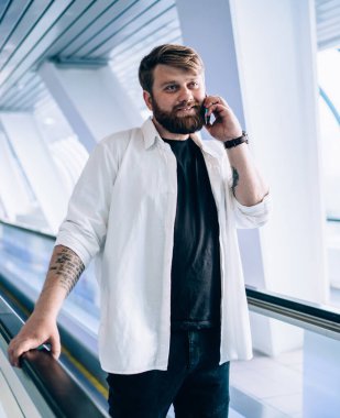 Elinde siyah tişört ve beyaz gömlekli şık sakallı adam cep telefonuyla konuşuyor hafif geometrik tünelde tırabzan tutuyor.