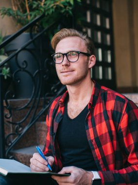 Gözlüklü, entellektüel kızıl saçlı erkek. Kırmızı ve siyah tişörtlü. Not defterine yazları granit merdivenlerde oturan.