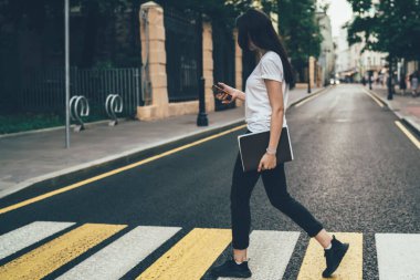 Yazın bulanık şehir manzarasına karşı yaya geçidinde yürürken ekranına odaklanan ve akıllı telefon kullanan günlük kıyafetlerle tanınamayan koyu renk saçlı kadının yan görüntüsü