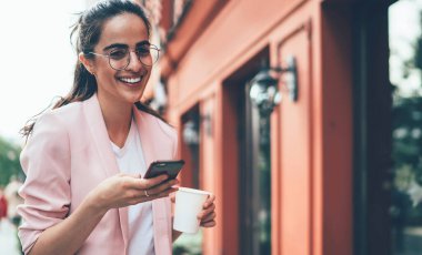 Düşük açılı, gözlüklü, sıcak içecekli zarif bir kadın cadde boyunca yürürken kameraya bakıp gülecek ve bulanık dış görünüşe karşı akıllı telefonuyla etkileşime girecek.