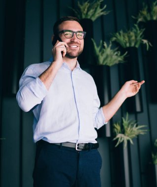 Modern ofisin koridorunda saksı bitkileriyle akıllı telefon konuşmaları yaparken gülümsemenin ve el kol hareketleri yapmanın düşük açısı.