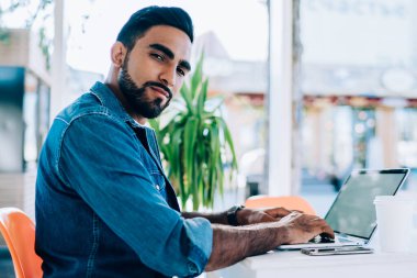 Dizüstü bilgisayar kullanarak yaz kafeteryasında oturup kameralara bakarak proje üzerinde çalışan serbest giyinen sakin etnik bir vatandaş.