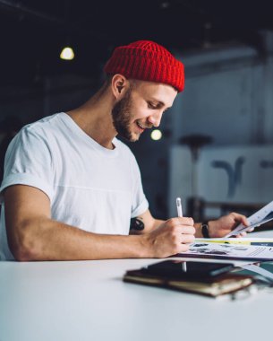 Kırmızı şapkalı genç adam, elinde akıllı telefon tableti defteriyle masada oturuyor ve iş sırasında gazetelere notlar yazıyor.
