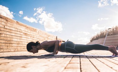 Gün ışığında dar tahta patikada dinlenirken rahat minder üzerinde yoga yapan kaslı, narin kadının yan görüntüsü