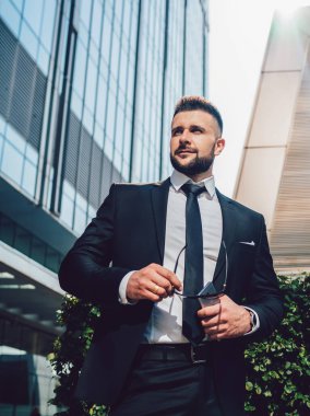 Düşük açılı, koyu renk takım elbiseli patron güneş gözlüğü ve akıllı telefon taşıyor ve şehir caddesindeki modern binaların dışındaki çalıların yanında dikilirken yüzünü çeviriyor.