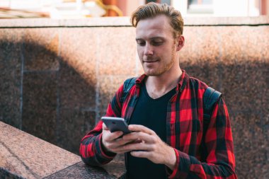 İşaretli kırmızı ve siyah tişörtlü odaklanmış gezgin cep telefonu izliyor binanın karşısında duruyor ve şehirdeki granit parmaklıklara yaslanıyor.