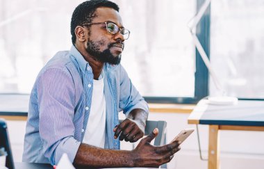 Orta yaşlı Afrikalı Amerikalı adam çalışırken bilgisayar ve kağıtlarla masada otururken akıllı telefonu elinde tutuyor ve düşünüyor.