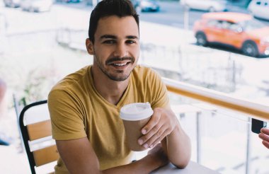 Gündelik kıyafetlerle gezen mutlu bir adam kafeteryada kameraya bakarak bir fincan kahve içen bir adamla iletişim kuruyor.