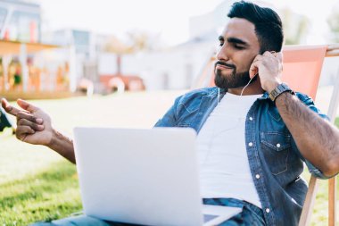 Rahat rahat gülümseyen, sıradan kot gömlekli, kulaklıklı, dizüstü bilgisayarlı müzik dinleyen, güneşli bir parkta vakit geçiren genç bir erkek.