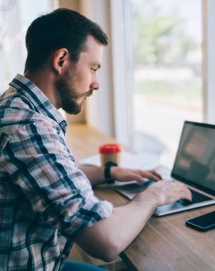 Gömleği kollu, odaklanmış sakallı bir adam bir fincan kahve alıp kafeteryada bilgisayarla uzaktan kumandayla çalışıyor.