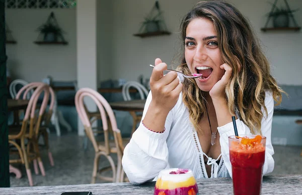 Attractive happy woman in light white blouse eating with pleasure delicious yogurt dessert and drinking smoothie while sitting in outdoor cafe looking at camera