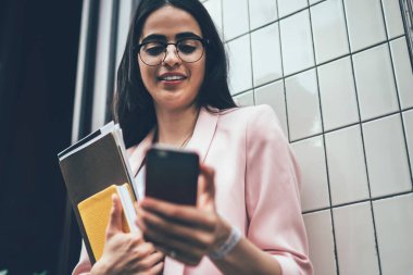 Elinde kağıtlar ve günlükle bina duvarının yanında duran ve internette cep telefonundan haber okuyan başarılı, modern meşgul bir kadından.