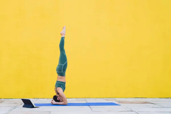 Side view of sportive slim woman in green outfit doing tripod headstand with legs raised straight up on yoga mat near tablet outdoors