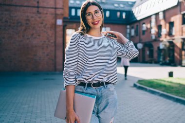 Üniversite kampüsünde modern dizüstü bilgisayarı olan mutlu kız öğrencinin yarım boy portresi, kameraya gülümseyen gözler için optik gözlüklü pozitif hipster kız.