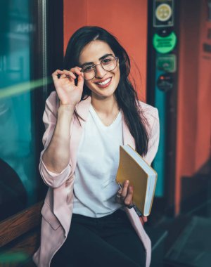 Yüksek açılı, zarif elbiseli, not defteri takan, gözlüklü ve şehirde ahşap bankta dinlenirken kameraya gülümseyen hoş bir yönetici kadın.