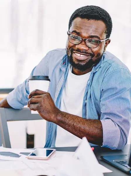 Content adult black man in glasses holding cup of coffee in hand and smiling at camera while sitting at laptop