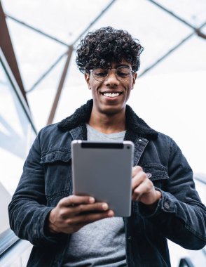 Optik gözlüklü neşeli Latin adam yürüyen merdivende aşağı iniyor ve dokunmatik ped aracılığıyla online sohbetten heyecanlanıyor, milenyum pozitif erkek blogcu mesajlar alıyor