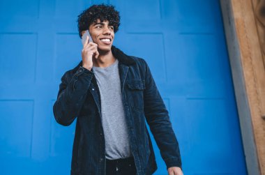 Gülümseyen, siyah ceketli ve kot pantolonlu genç, kıvırcık etnik adam caddede mavi şehrin kapısının önünde dikilirken telefonla konuşuyor.