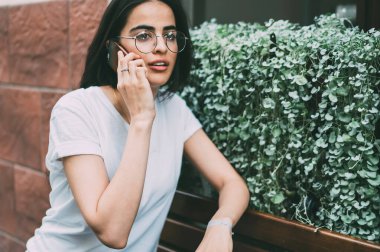 Kendine güveni tam genç bir kadın, gündelik giysiler ve ahşap bankta eli olan gözlüklü, akıllı telefon kullanarak kafenin dışında oturan bir arkadaşla konuşuyor.