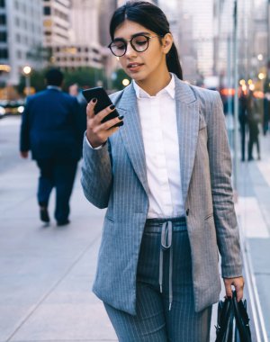 Resmi kıyafetli, gözlüklü, siyah saçlı, siyah çantalı genç bir kadın sokakta yürürken insanların arka planında geziniyor.