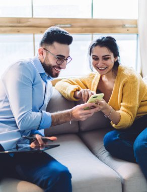 Resmi tablet ve cep telefonuyla genç etnik işadamı ve sıradan bir kadınla koltukta oturup akıllı telefondan izliyor ve evde gülüyor.