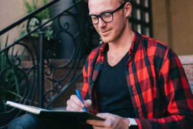 Neşeli, kızıl saçlı, gözlüklü, işaretli kırmızı ve siyah tişörtlü bir erkek. Yazın şehirde granit merdivenlerde oturan kalemle not alıyor.