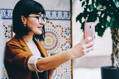 Keyifli uzaktan kumandalı bir işçinin sosyal medya için akıllı telefon kullanarak kafede oturarak selfie çekmesinin yan görüntüsü.