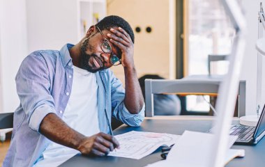 Yorgun sakallı siyah adam alnına dokunuyor ve masa başında oturmuş, ofis planları üzerinde çalışırken kameraya bakıyor.