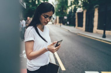 Gözlüklü ve günlük giysileri olan, ekranda yoğunlaşan ve kaldırım kenarında bulanık sokaklara karşı kaldırım kenarında duran akıllı telefonuyla iletişim kuran sakin bir milenyum çalışanı.