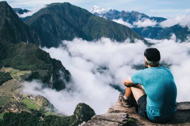 Aktif giyinmiş profesyonel bir adamın dağların zirvesinde dinlenmesi ve Machu Picchu 'nun yeşil tepelerine hayran olması ve yürüyüş sırasında doğal çevrenin nefes kesici manzarası.