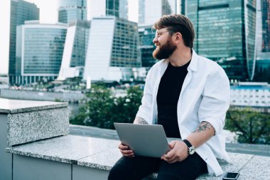 Kollarını sıvayıp dizüstü bilgisayar kullanarak rahatlayan beyaz gömlekli sıradan bir erkek kentsel arka plana bakıyor.