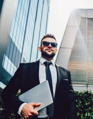 Karizmatik, kendine güvenen, güneş gözlüklü, şık siyah takım elbiseli, dizüstü bilgisayarı olan, caddede durup gülümseyen ve çağdaş yüksek mevkilere karşı kendi başarısının tadını çıkaran bir yönetici.