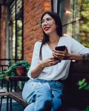 Yüzünde sevimli bir gülümsemeyle mutlu bir Latin kadın elinde dijital cep telefonu cihazıyla ve boş zamanlarında internetteki telefon konuşmaları için seviniyor. Akıllı telefonlu neşeli bir bayan milenyumu kafenin terasında oturuyor.