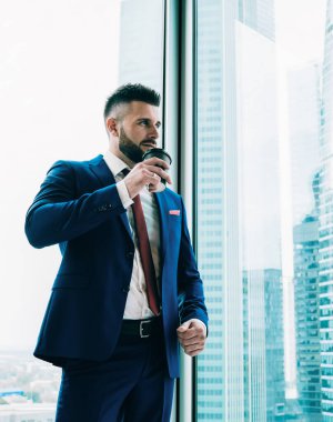 Gökdelendeki ofis odasında sıcak içecek içerken büyük pencereden dışarı bakan resmi takım elbiseli zeki, düşünceli zarif bir işçi.