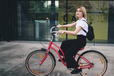 Kendine güvenen, gündelik kıyafetli, siyah sırt çantalı, bisiklet süren ve kameraya bakan modern sarışın kadının yan görüntüsü.