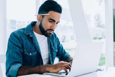 Çalışkan, düşünceli, günlük giysiler içinde, kulağının arkasında kalemle ekranda gezinen ve bilgisayarla etkileşime giren İspanyol asıllı serbest yazar açık havada beyaz terasta elinde not defteriyle otururken.