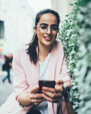 Modern şehrin kentsel sokaklarında kameralara bakarak arkadaşlarıyla akıllı telefon mesajları kullanan gündelik kıyafetleri ve gözlükleriyle hoş bir bayan.