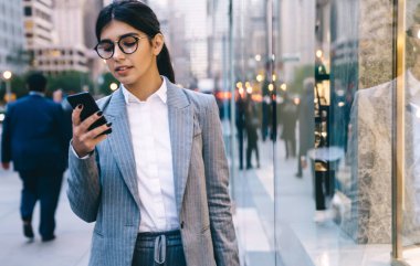 Resmi kıyafetli, gözlüklü, siyah saçlı, siyah çantalı genç bir kadın sokakta yürürken insanların arka planında geziniyor.