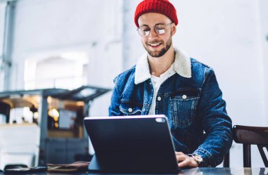 İnternette sörf yapmak ve bilgi aramak için dijital cihaz kullanan sıradan giyinmiş mutlu yakışıklı genç adam telefon ve bardakla çatı katında otururken...