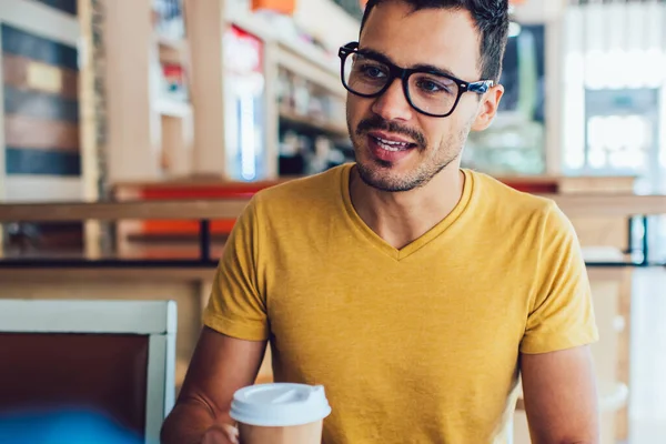Confident remote worker in casual clothes and glasses creating startup with crop partner discussing strategy taking hot drink sitting at table in cafe