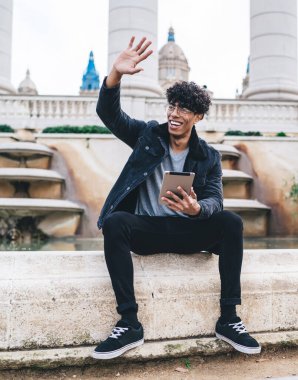 Siyah elbiseli genç Afro-Amerikan erkek öğrenci çeşmenin kenarında oturmuş, elinde tabletle kentin arka planında el sallıyor.