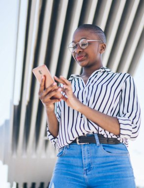Çizgili bluz ve kot pantolonlu şık, kel, Afro-Amerikan kadın yazın köprü altında dururken akıllı telefondan mesajları kontrol ediyor.