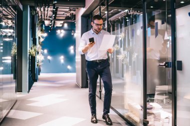 Ofis kıyafetleri içinde, ofis binasında cep telefonuyla dolaşırken dokümanlara bakan, gözlüklü, hoşnutsuz, sakallı yönetici erkek.